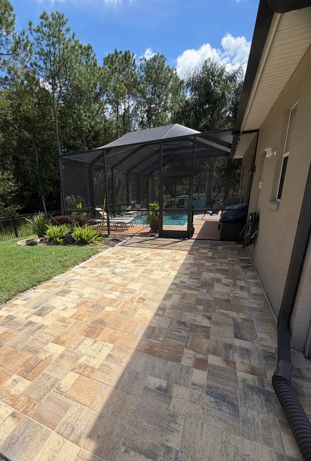 Paver pool deck and adjacent patio area next to a home with a screened pool enclosure and outdoor furniture on a sunny day.