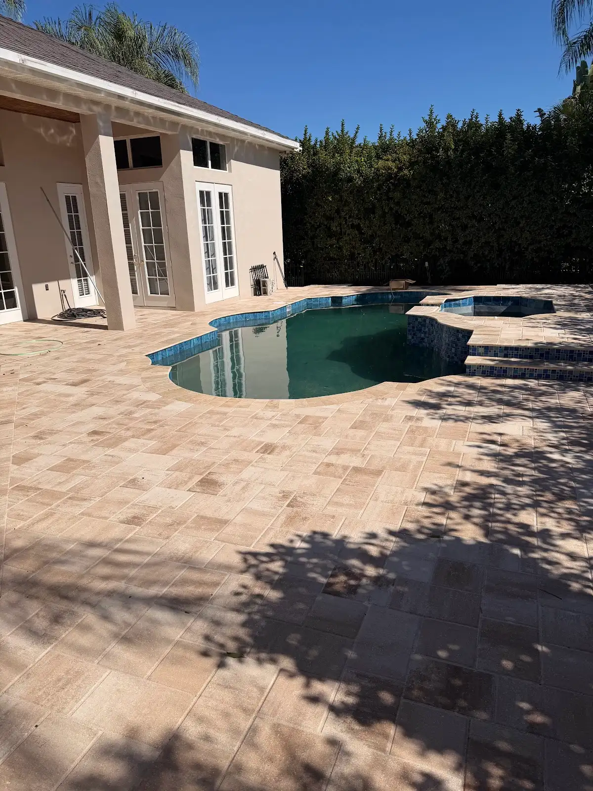 A backyard pool area featuring a clean, newly installed beige paver pool deck surrounding a pool and spa, shaded by nearby trees.