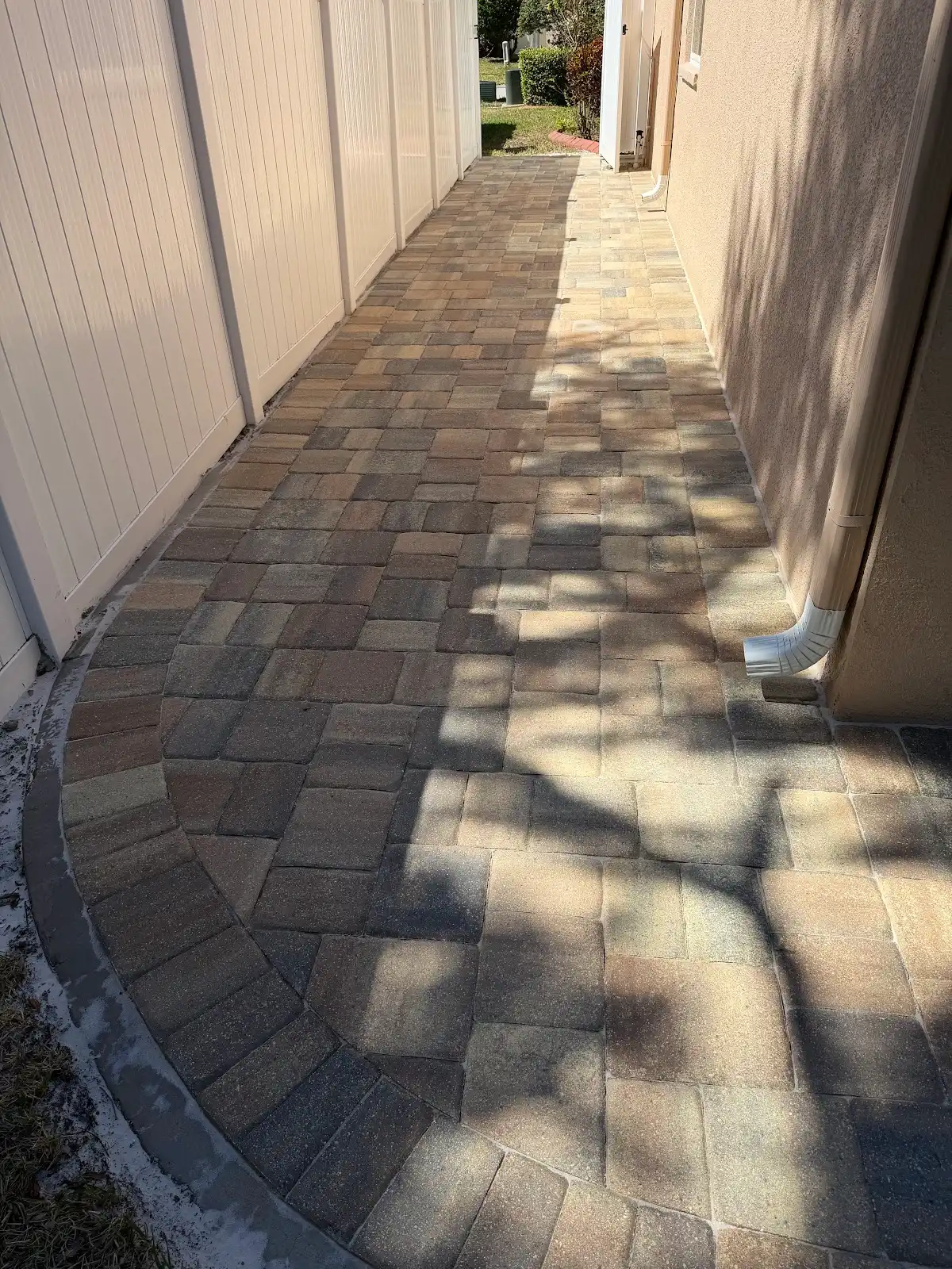 Curved walkway made of multi-colored pavers between a white fence and beige house wall with gutter downspout