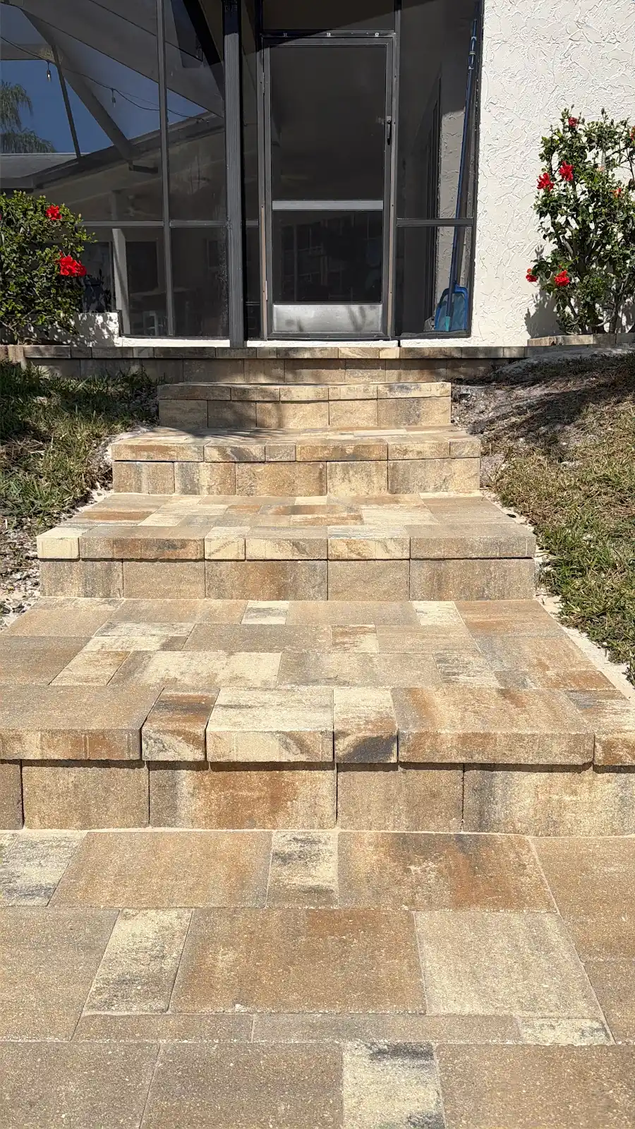 Multi-level beige and tan paver steps and walkway leading to a screened porch door with plants and shrubs on sides.