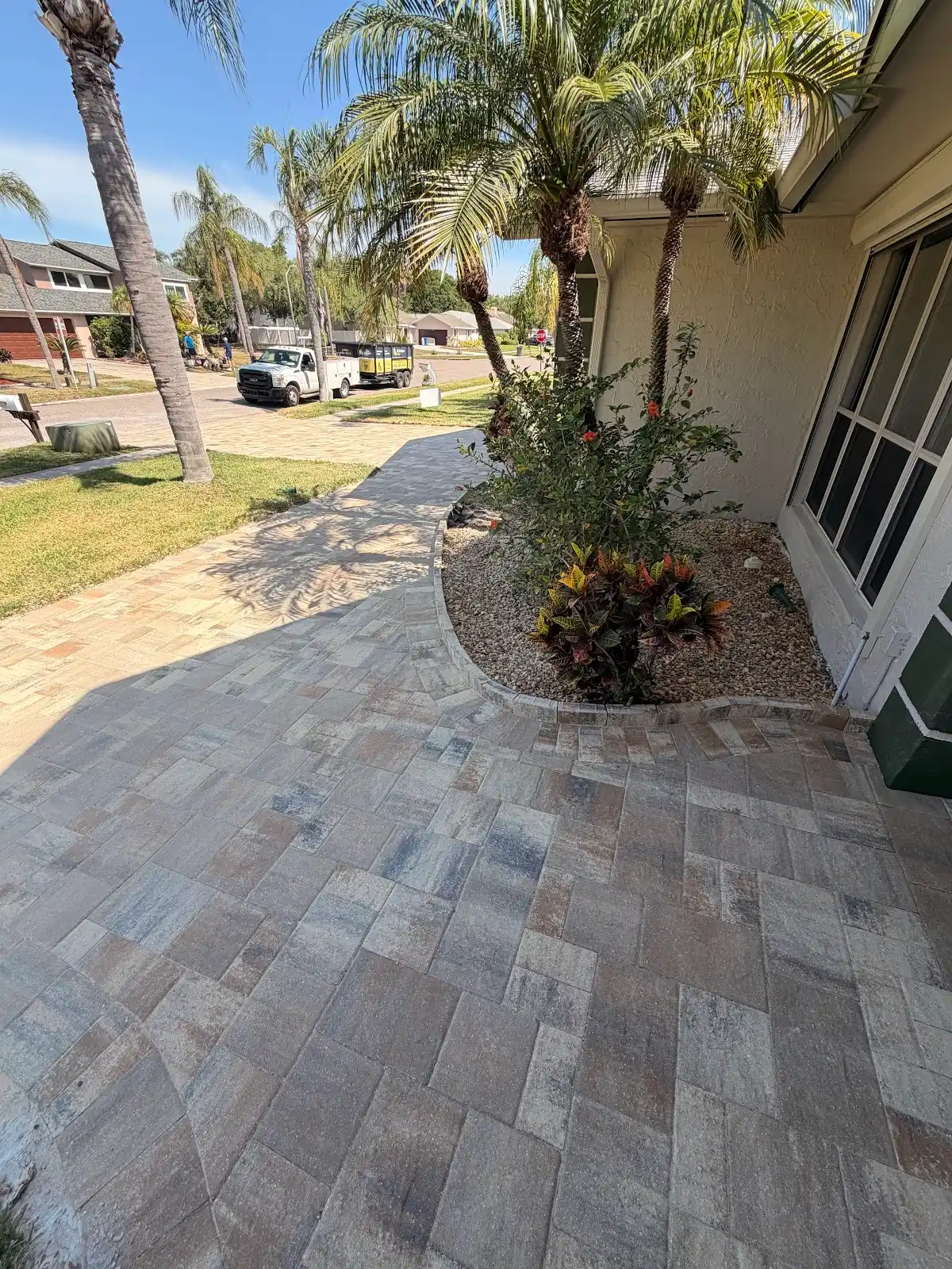 A finished paver walkway and driveway next to a house, with palm trees and plants in landscaped beds under a clear sky.