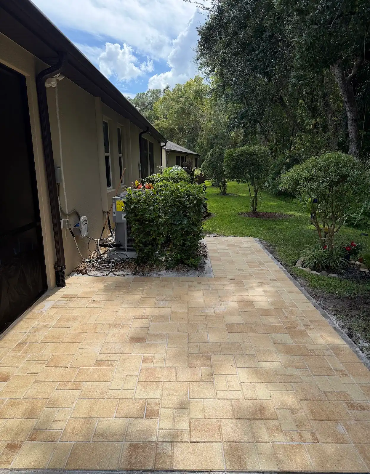 Newly installed light tan paver walkway along side of a beige house, bordered by green bushes and grass under a partly cloudy sky.