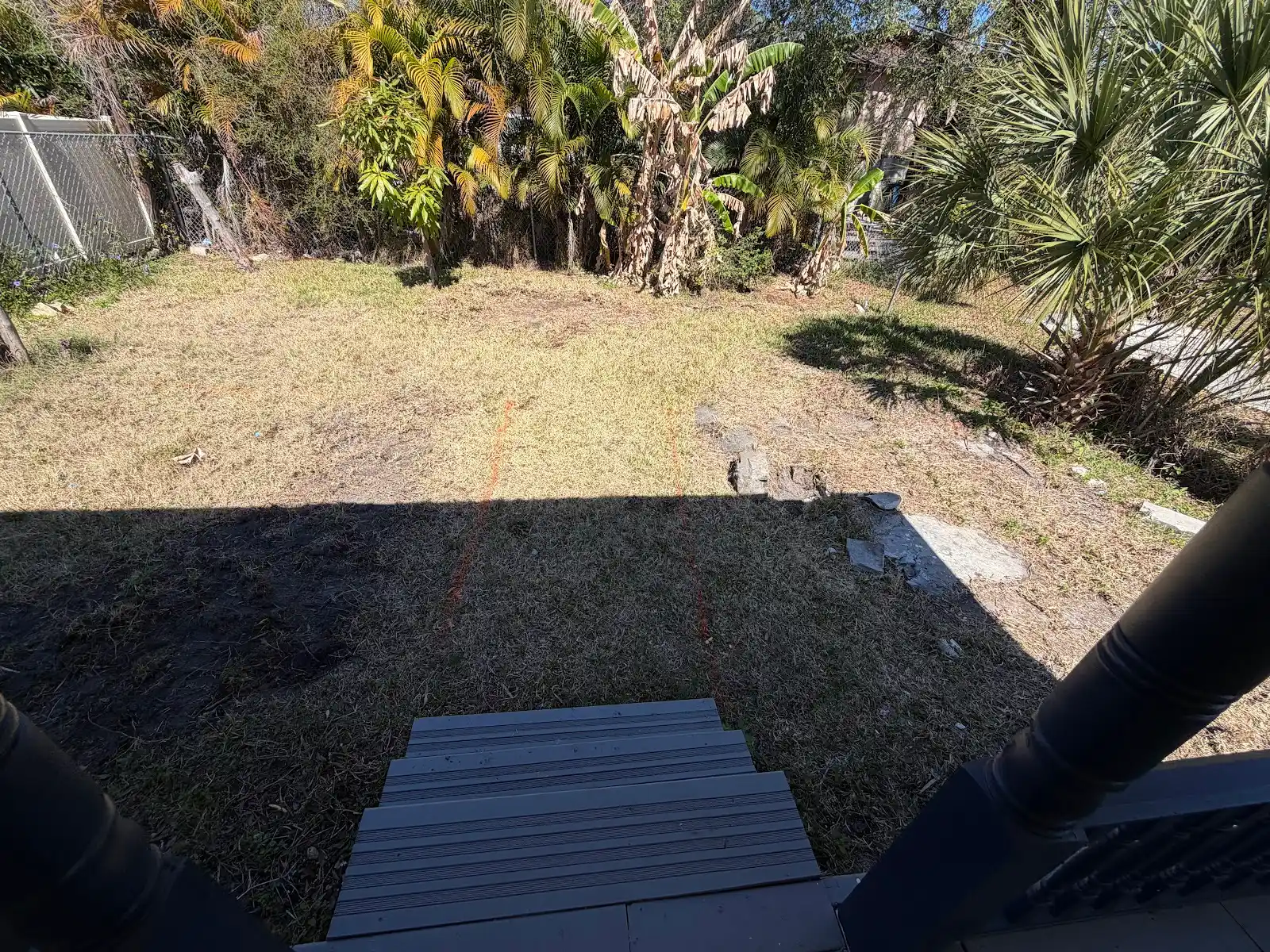 View of a backyard with dry grass and orange markings on the ground, seen from a porch with steps and black railing.