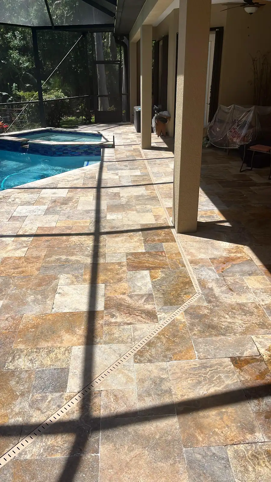 View of a finished travertine marble pool deck and patio area with beige and earth tone tiles next to a pool and spa, under a screened enclosure on a sunny day.