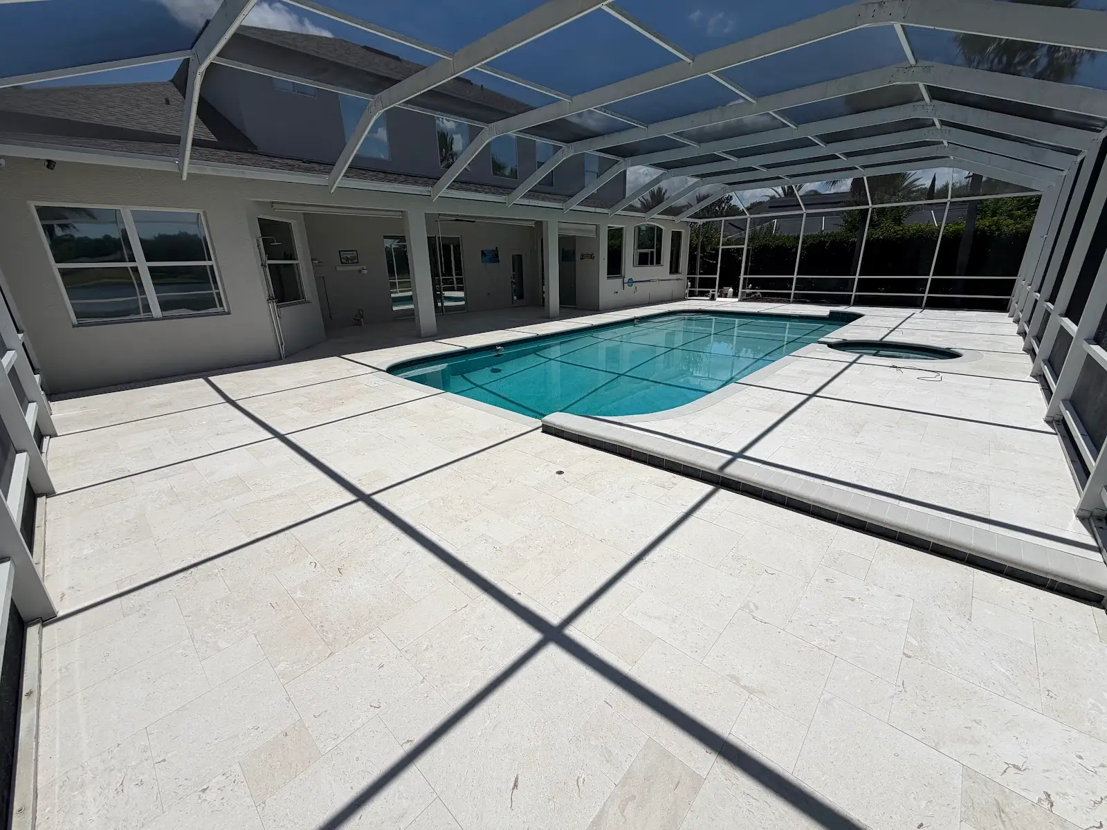 Light beige travertine marble tiles surrounding a rectangular blue pool and spa inside a screened patio enclosure under a clear sky.