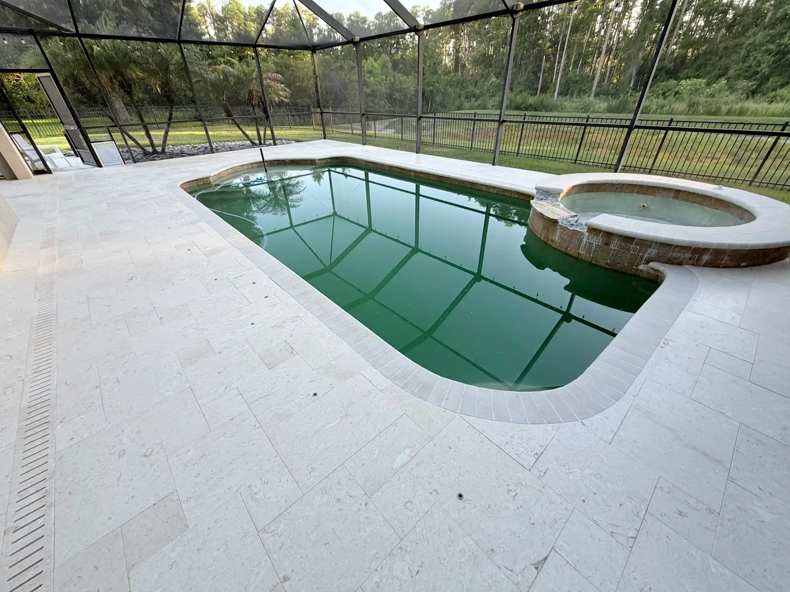 Light beige travertine marble pool deck and spa surrounding a swimming pool with screened enclosure and wooded background