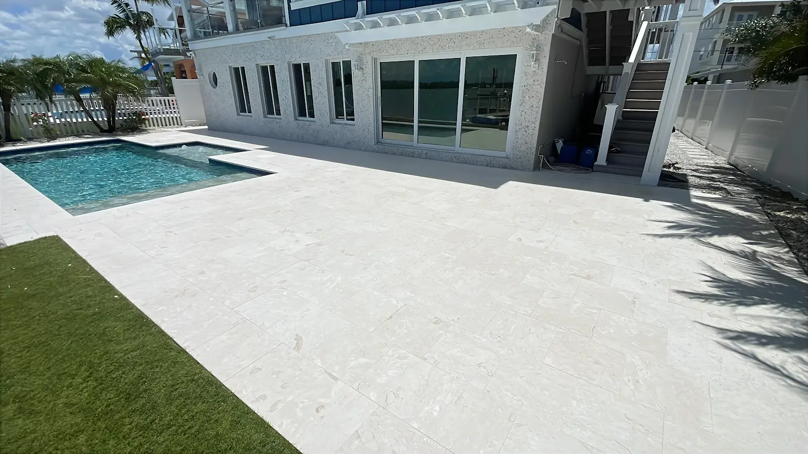 Light-colored travertine marble pool deck next to a swimming pool and artificial turf lawn outside a residential house in Tampa Bay