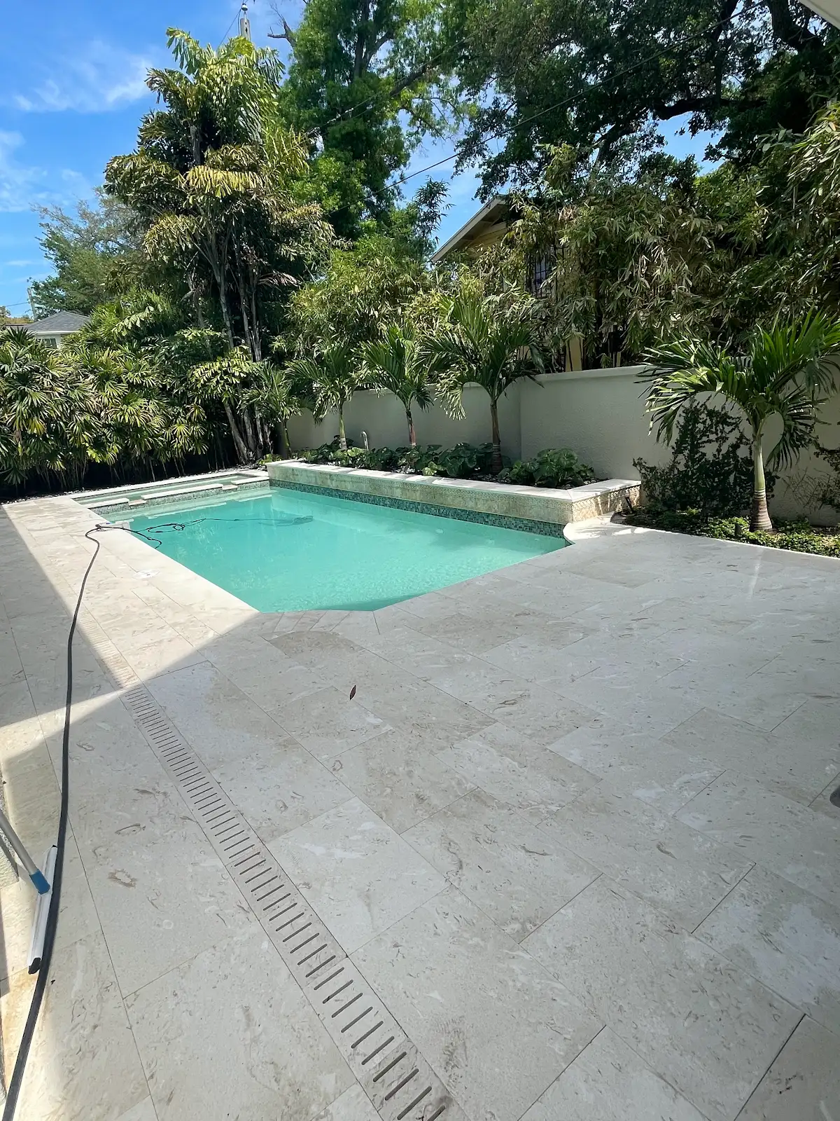 Rectangular swimming pool with clear blue water surrounded by a travertine marble tiled pool deck and lush greenery in the background.