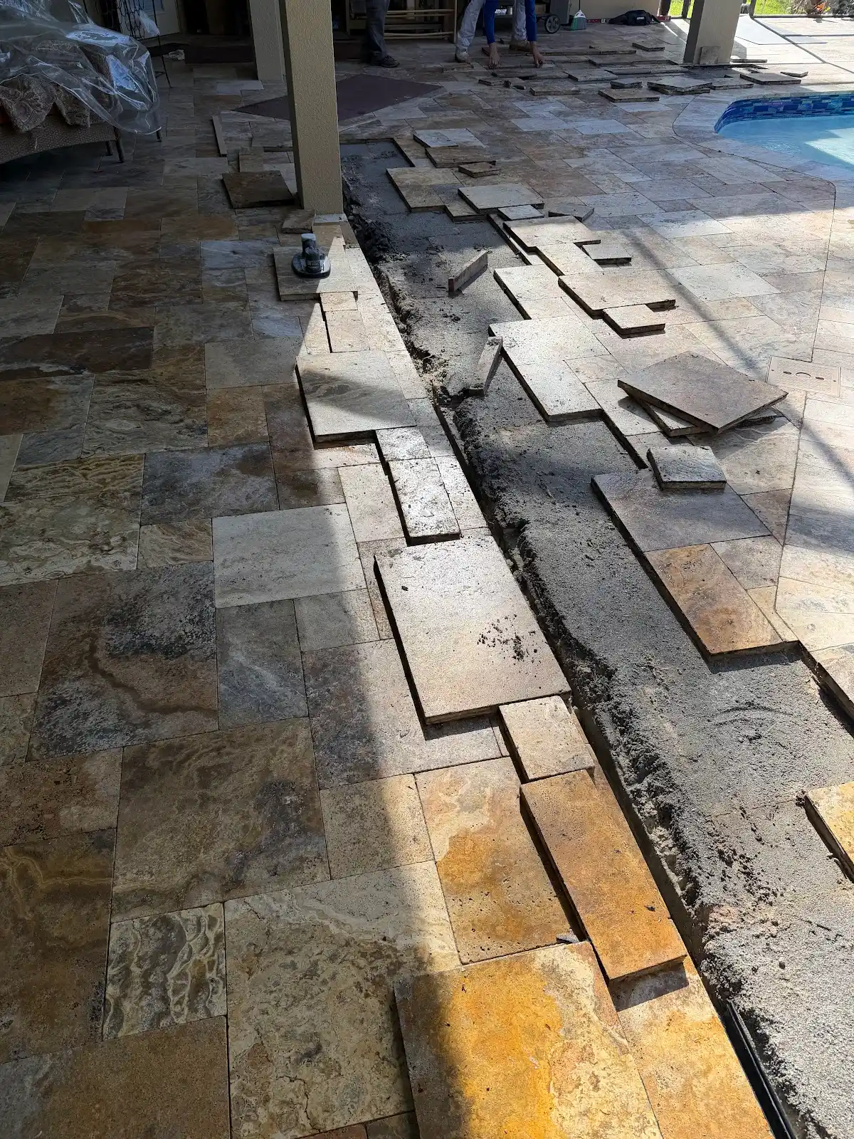In-progress installation of travertine marble tiles around a pool deck with tiles partially removed and new tiles being placed.