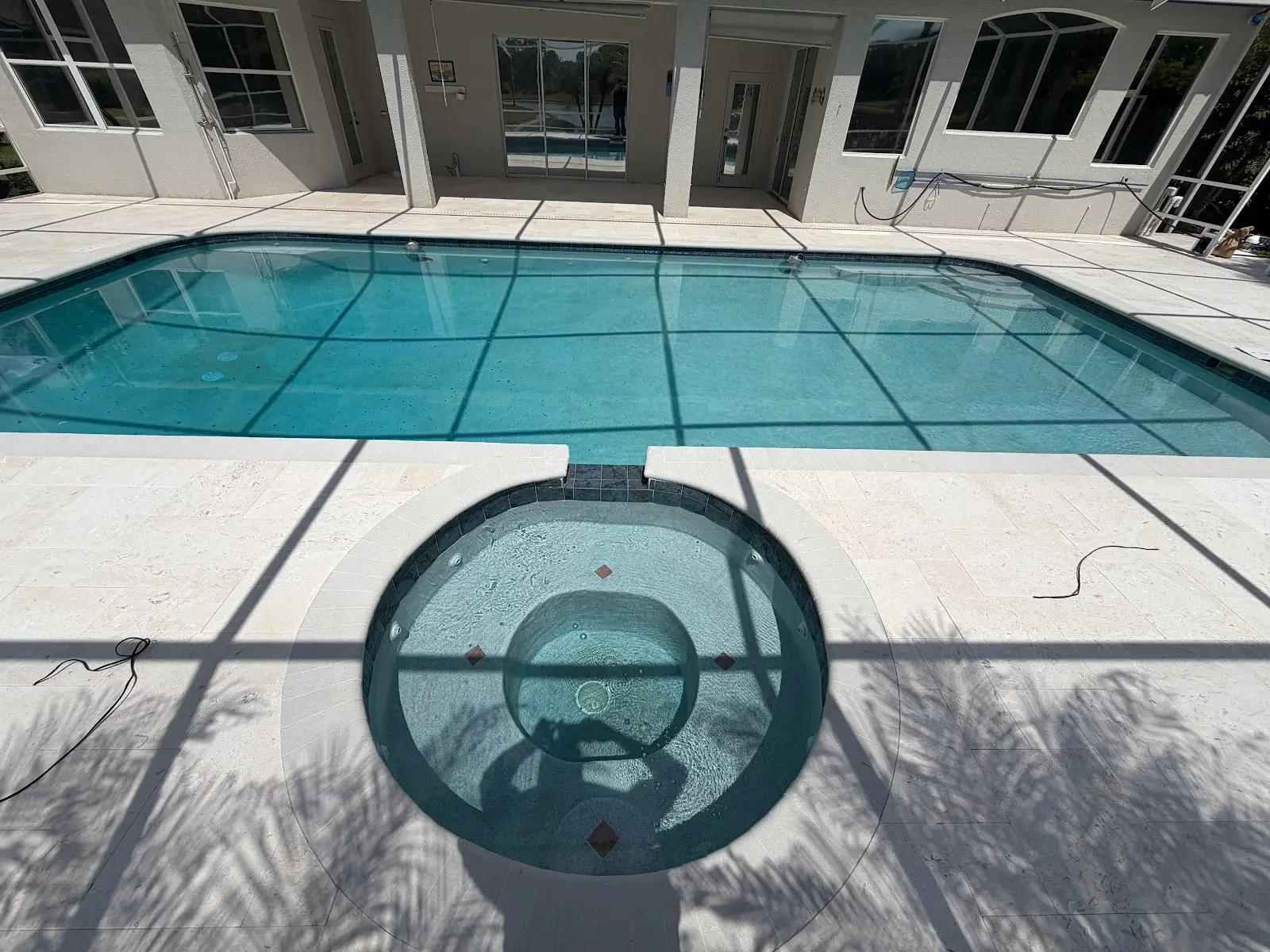 Clean travertine marble pool deck and spa area with clear blue swimming pool and surrounding house exterior in sunlight