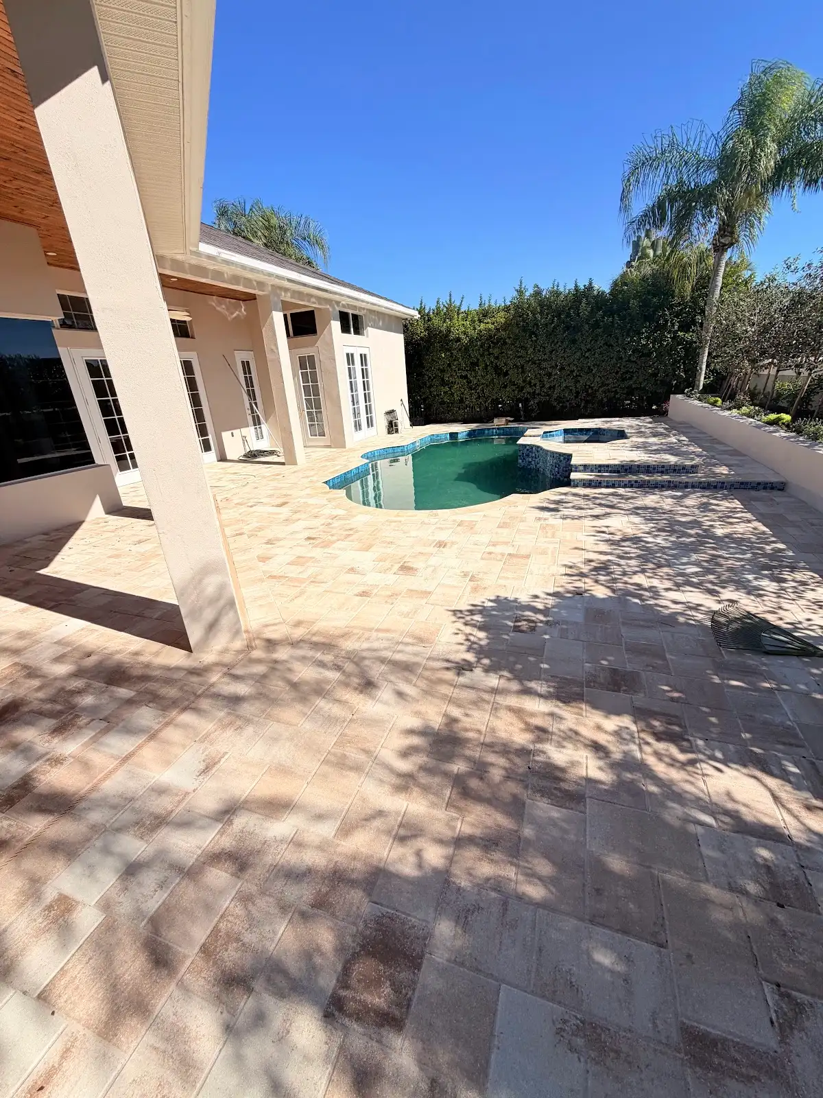 A clear view of a light beige travertine marble pool deck surrounding a swimming pool next to a house with beige walls and white doors, under a blue sky