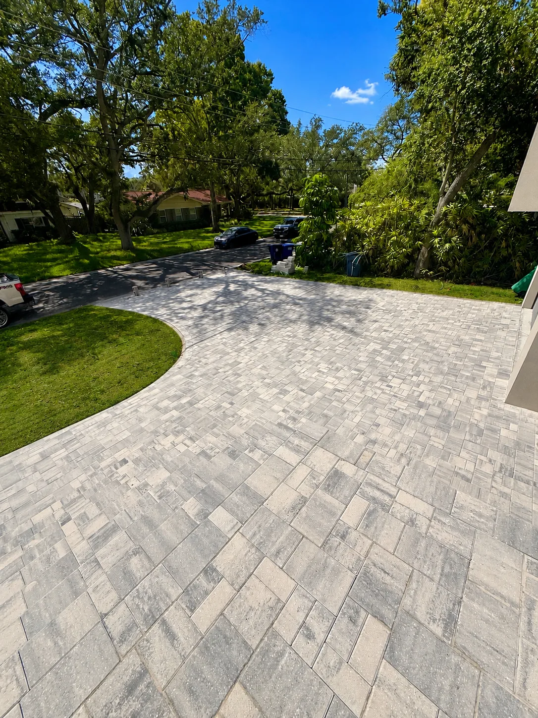 Completed curved gray paver driveway bordered by lawn and mature tropical landscaping