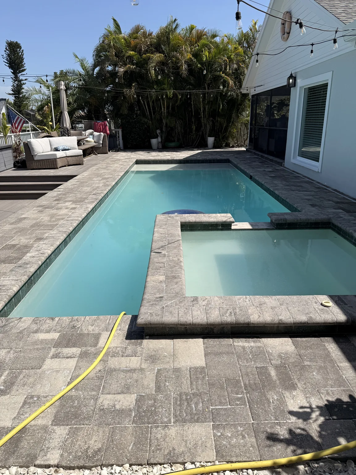 Completed gray paver pool deck surrounding a rectangular swimming pool beside a home